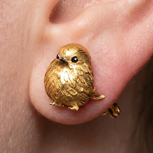 May include: A gold-toned earring shaped like a small, round bird with textured plumage and dark, prominent eyes. The bird figure is depicted in a seated position with detailed wing and feather carvings, secured to the earlobe with a metal clip mechanism.