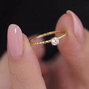 May include: A thin gold ring featuring a textured, hammered band and a single, round, clear faceted gemstone set in a four-prong setting. The band has a distinct, repeating indented pattern along its entire circumference, providing a reflective surface.