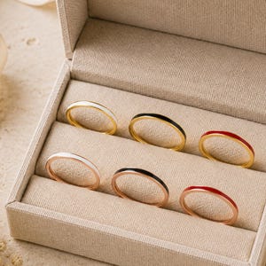 May include: A collection of six thin metal rings arranged in a beige jewellery box. The top row features rings with white, black, and red enamel inlay on gold bands. The bottom row features rings with white, black, and red enamel on rose gold bands.