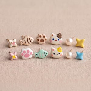 May include: A collection of twelve small stud earrings featuring various shapes including white and pink cat paws, light blue and brown fish, white cat faces with colored spots, white pearls, and four-pointed stars in yellow, blue, and pink colors.