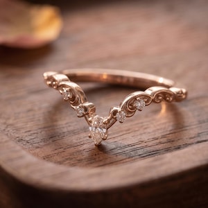 May include: A rose gold V-shaped ring featuring a central pear-cut diamond and smaller round-cut diamonds set along the band. The metal band displays intricate scrollwork detailing that follows the chevron contour of the ring design.