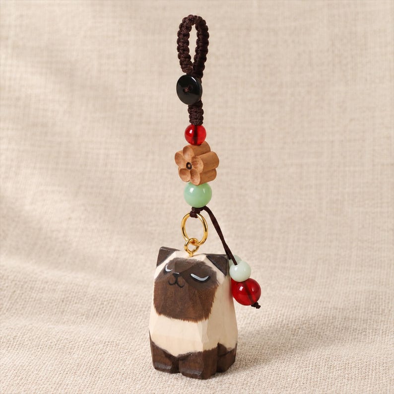 May include: A decorative hanging charm featuring a carved wooden Siamese cat figure with dark brown and cream colouring. The charm is suspended by a brown braided cord adorned with a black bead, a red bead, a light green bead, and a wooden flower.