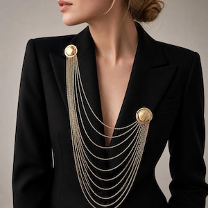 May include: A black blazer featuring a gold-toned brooch accessory. The brooch consists of two circular, polished gold discs connected by multiple draped gold chains that hang in a cascading, layered pattern across the front of the garment.