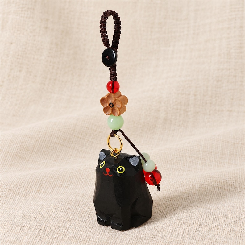 May include: A black cat charm carved from wood with yellow eyes and a red mouth, suspended from a brown braided cord. The cord features a black button, a red bead, a light brown wooden flower, and a pale green bead, with additional beads.