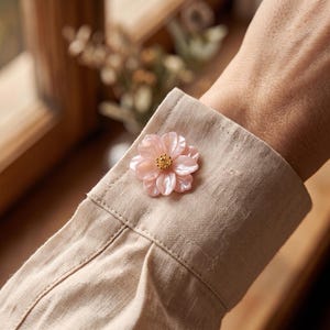 Peut inclure: Un bouton en forme de fleur rose clair avec une finition nacrée et un petit centre doré, fixé sur la manchette d'une chemise à manches longues en lin beige. Le bouton présente des pétales superposés et un détail d'étamines.