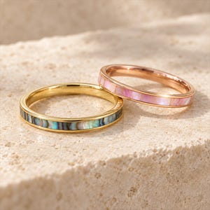 May include: Two thin metal rings. One gold-toned ring features an inlay of iridescent dark blue and green abalone shell. The second rose gold-toned ring features an inlay of iridescent pink shell. Both bands have a smooth, polished finish.