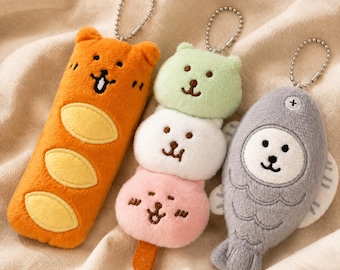 May include: Three plush keychains featuring an orange bear-shaped baguette, a stack of three pastel-coloured animal-faced dango balls on a stick, and a grey fish-shaped character with a white face, each attached to a silver metal ball chain.