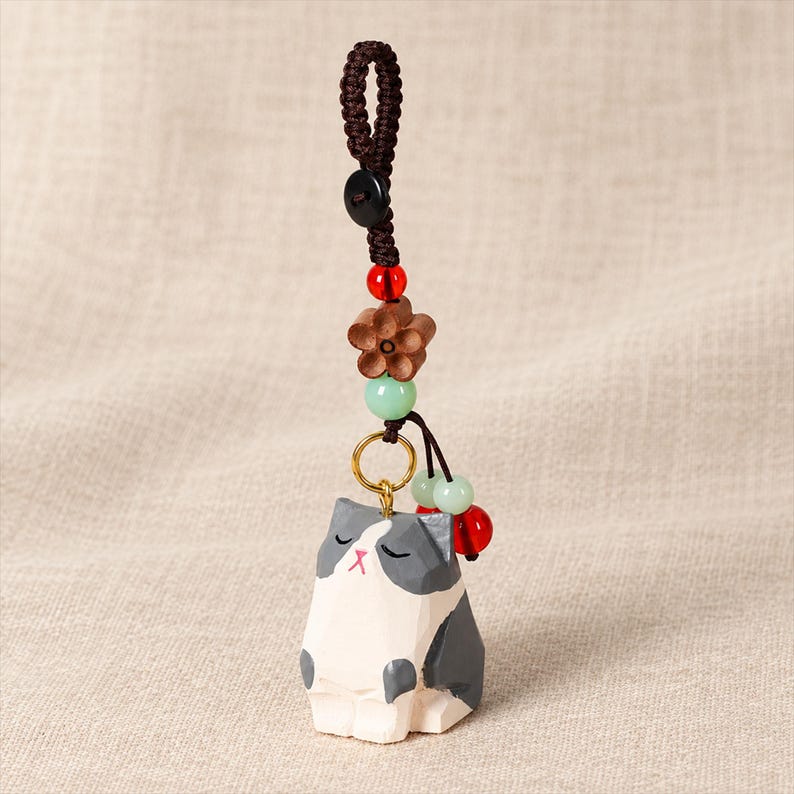 May include: A decorative keyring featuring a hand-carved wooden cat figurine with grey and white painted details. The cat is attached to a brown braided cord with a small black button, a wooden flower bead, and several small red and light green beads.