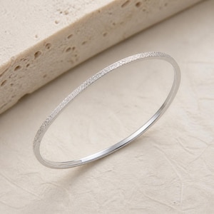 May include: A thin, circular bangle bracelet with a textured, sparkling silver-toned finish. The band features a consistent, fine-grained surface that catches the light, creating a subtle shimmer across the entire circumference of the jewelry piece.