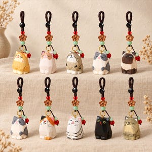 May include: Ten hand-carved wooden cat charms arranged in two rows. Each cat features a unique coat pattern and expression, suspended from a dark brown braided cord with a wooden flower bead, a small green bead, and a red bead accent.