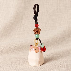 May include: A hand-carved wooden cat charm featuring a cream-coloured body with brown facial markings and pink ears. The charm hangs from a dark brown braided cord adorned with a wooden flower bead, a small black button, and red and light green beads.