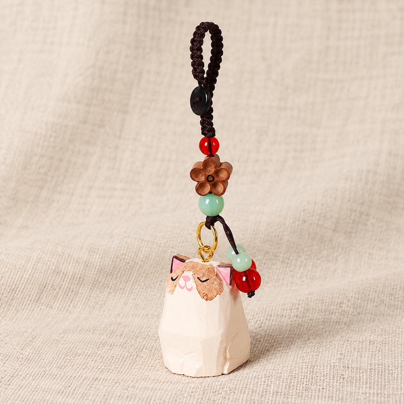 May include: A hand-carved wooden cat charm featuring a cream-coloured body with brown facial markings and pink ears. The charm hangs from a dark brown braided cord adorned with a wooden flower bead, a small black button, and red and light green beads.