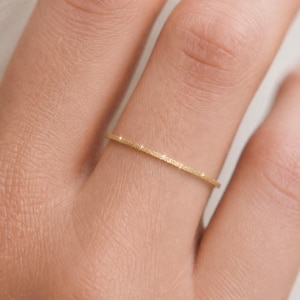 May include: A thin, yellow gold ring with a textured, matte finish. The band is narrow and circular, featuring a consistent surface pattern that catches the light at several points along its circumference, creating a subtle sparkle on the metal.
