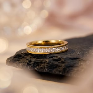 May include: A gold band ring featuring a continuous channel setting of square-cut clear stones that wrap around the entire circumference of the metal band, creating a uniform and reflective surface across the exterior of the jewelry piece.