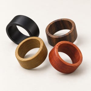 May include: Four wide wooden rings arranged in a cluster. The collection includes one solid black ring, one dark brown ring with visible grain, one light tan ring, and one reddish-orange ring. Each ring features a smooth, rounded exterior surface.