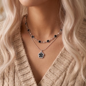 Peut inclure: Un ensemble de colliers superposés en argent composé d'une chaîne supérieure avec de petites pierres circulaires noires et de minuscules perles blanches, et d'une chaîne inférieure avec un pendentif fleur en argent au centre noir.