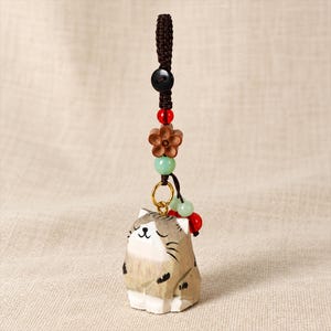 May include: A carved wooden cat charm featuring a light brown and white striped pattern with closed eyes and whiskers. The charm hangs from a dark brown braided cord adorned with a black button, a red bead, a wooden flower, and light green beads.