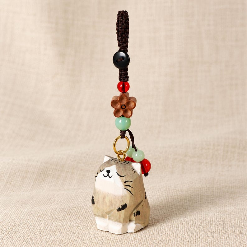 May include: A carved wooden cat charm featuring a light brown and white striped pattern with closed eyes and whiskers. The charm hangs from a dark brown braided cord adorned with a black button, a red bead, a wooden flower, and light green beads.