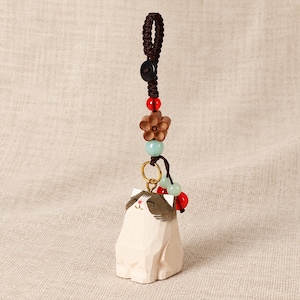 May include: A carved wooden cat charm featuring a white body with brown markings on the head, suspended from a dark brown braided cord. The charm includes a small wooden flower bead, a red bead, and light green beads attached to a gold ring.