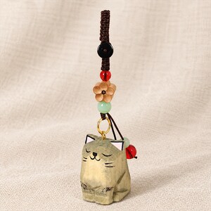 May include: A wooden cat charm featuring a carved, light-brown feline figure with closed eyes and pink-tipped ears. The charm hangs from a brown braided cord adorned with a black bead, a red bead, a light-brown wooden flower, and a pale green bead.