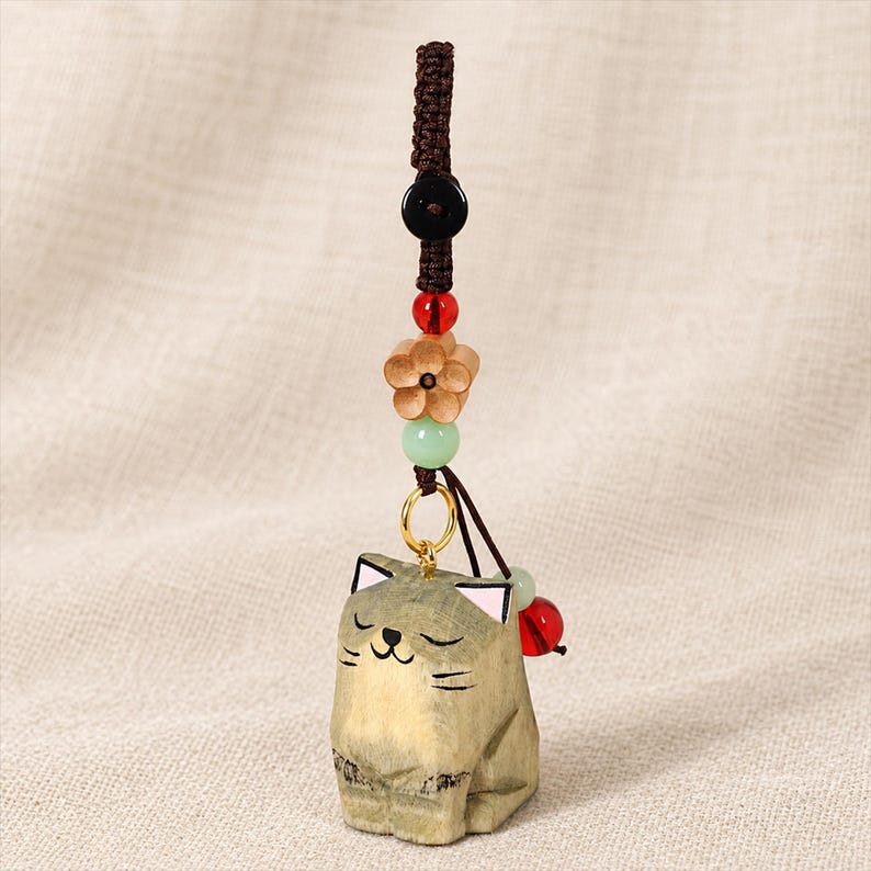 May include: A wooden cat charm featuring a carved, light-brown feline figure with closed eyes and pink-tipped ears. The charm hangs from a brown braided cord adorned with a black bead, a red bead, a light-brown wooden flower, and a pale green bead.