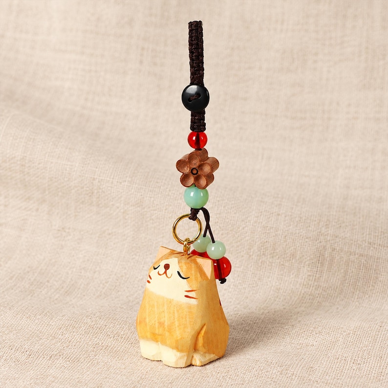 May include: A carved wooden cat charm featuring a light orange and cream finish with painted facial details. The charm hangs from a dark brown braided cord adorned with a black button, a small wooden flower, and red and light green beads.