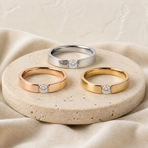 May include: Three rings featuring a tension-set round brilliant-cut clear gemstone. One ring is silver-toned, one is rose gold-toned, and one is yellow gold-toned. Each band has a smooth, polished finish and a flat profile with a central stone.