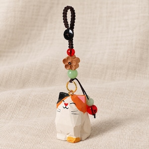 May include: A calico cat charm featuring a faceted white, orange, and black wooden body with a heart-shaped nose. The charm hangs from a dark brown braided cord adorned with a black button, a red bead, a wooden flower, and a light green bead.