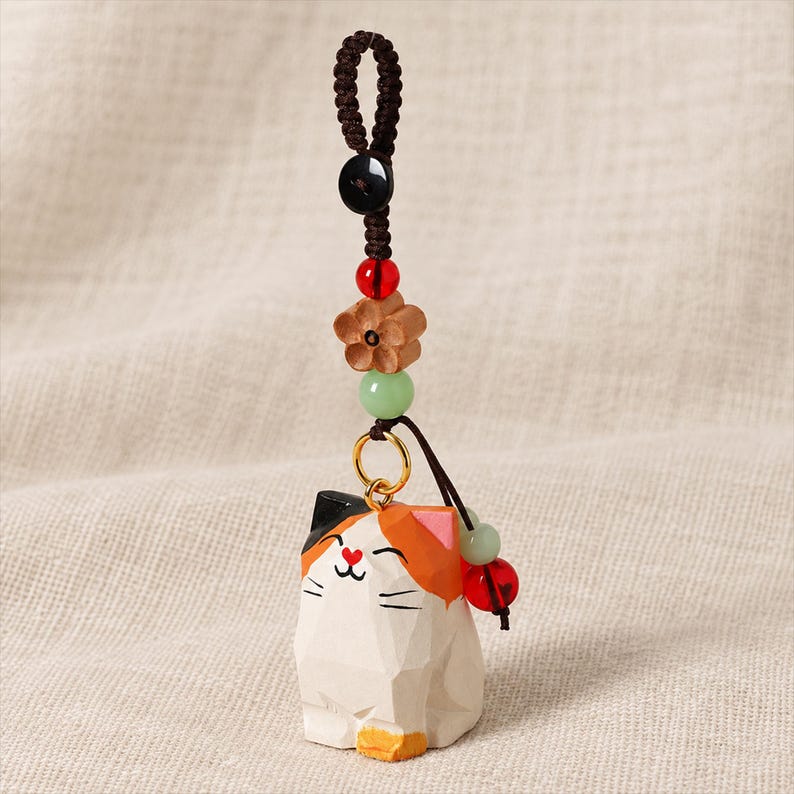 May include: A calico cat charm featuring a faceted white, orange, and black wooden body with a heart-shaped nose. The charm hangs from a dark brown braided cord adorned with a black button, a red bead, a wooden flower, and a light green bead.