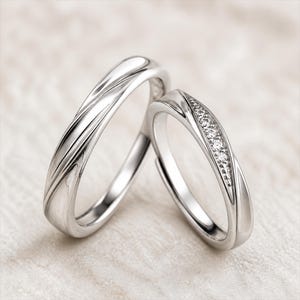 May include: Two silver-toned rings with a polished finish. One ring features a smooth, twisted metal band design. The second ring displays a similar twisted metal band with a row of five small, clear, round-cut stones embedded along the center.
