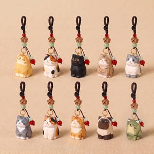 May include: Ten small, hand-carved wooden cat charms arranged in two rows. Each cat features a unique coat pattern and colour, including orange, calico, black, tabby, and grey. Each charm hangs from a dark cord with a small wooden flower and red bead.