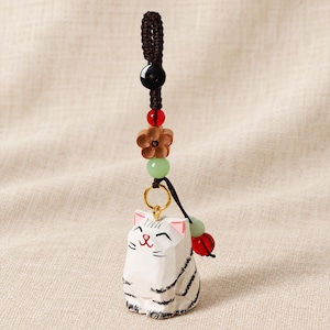 May include: A white carved wooden cat charm with black stripes and pink ears, suspended from a dark brown braided cord. The cord features a black bead, a red bead, a wooden flower-shaped bead, and a light green bead, with a gold ring connector.