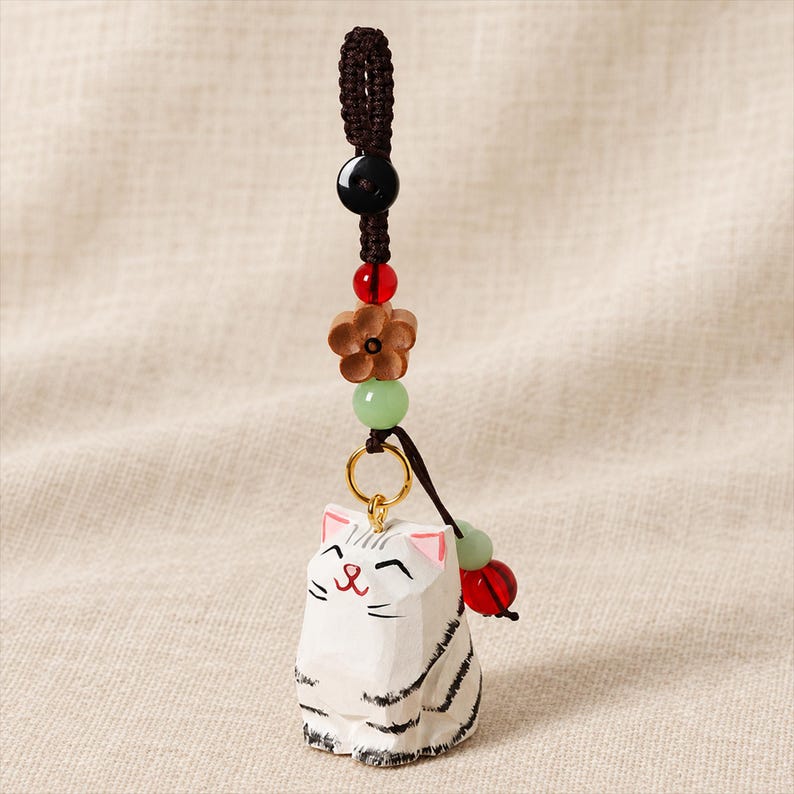 May include: A white carved wooden cat charm with black stripes and pink ears, suspended from a dark brown braided cord. The cord features a black bead, a red bead, a wooden flower-shaped bead, and a light green bead, with a gold ring connector.