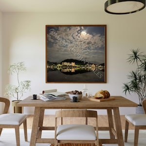 May include: A dining room scene featuring a wooden table with chairs, a framed landscape print, and decorative plants. The print depicts a nighttime scene with a body of water reflecting lights. The room has a neutral color palette.
