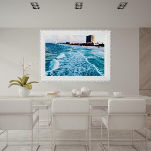 Könnte beinhalten: Ein gerahmtes Strandbild mit weißem Rahmen hängt über einem weißen Esstisch. Das Kunstwerk zeigt einen lebendigen blauen Ozean mit Wellen, einen Sandstrand und Gebäude in der Ferne. Weiße Stühle umgeben den Tisch.