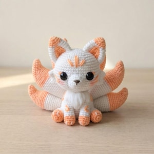 May include: A handmade crochet fox doll with white and orange details. The fox has large black eyes, a fluffy white chest, and a set of orange and white tails. The fox is sitting on a wooden surface.
