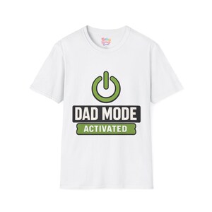 May include: A white short-sleeved t-shirt featuring a green power button icon above a black rectangular graphic with the white text DAD MODE and a green rectangular graphic below it with the white text ACTIVATED. The inner neck label reads Baby Licking Good.
