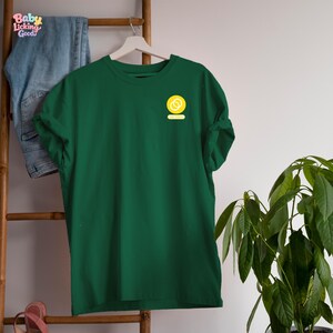May include: A dark green short-sleeved t-shirt featuring a small yellow circular logo on the left chest. The logo contains two interlocking white rings and the text "Baby Licking Good" in a stylized font. The shirt is displayed on a white hanger.