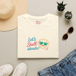 Può includere: Una t-shirt color crema con la grafica di una conchiglia che indossa occhiali da sole verdi e il testo Let's Shell abrate! in blu e rosa. La maglietta presenta uno scollo rotondo e un piccolo logo del marchio stampato all'interno del colletto.