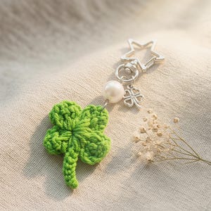May include: A green crocheted four-leaf clover keychain with a silver star-shaped clasp and a pearl bead. A small silver four-leaf clover charm hangs below the pearl. The keychain is set against a neutral fabric background.
