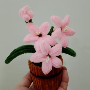 May include: A handmade craft featuring pink flowers with white stamens and green leaves, arranged in a brown pot. The flowers are made of a soft, fuzzy material, creating a whimsical floral arrangement.