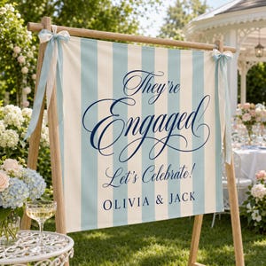 May include: A rectangular fabric sign featuring vertical light blue and white stripes, suspended from a wooden A-frame stand with light blue ribbons. The text reads "They're Engaged Let's Celebrate! OLIVIA & JACK" in a dark blue script and serif font.