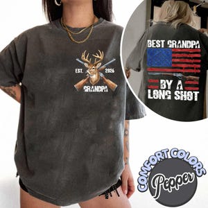 Puede incluir: Camiseta gris oscuro con una cabeza de ciervo, rifles cruzados y la palabra "GRANDPA" impresa en la parte delantera. La parte trasera de la camiseta presenta un diseño de bandera estadounidense con el texto "BEST GRANDPA BY A LONG SHOT".