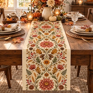 May include: A cream-colored table runner featuring a repeating botanical pattern of orange and red dahlias, brown oak leaves, acorns, wheat stalks, and green fern fronds, printed in a symmetrical, flowing vine-like arrangement.