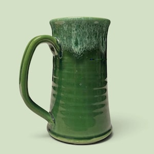 May include: A tall ceramic mug with a glossy forest green glaze and a textured, rippled body. The rim features a decorative drip effect with a lighter, mottled white and pale green finish that cascades down the upper portion of the vessel.