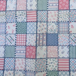 May include: A fabric textile featuring a patchwork design composed of square segments. The pattern includes various floral prints, stripes, and checkered motifs in shades of pink, blue, green, and white, joined by dark blue cross-stitch embroidery.