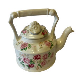 May include: A cream-colored ceramic teapot featuring a floral pattern of pink and red roses with green leaves. The teapot has a curved handle, a spout, and a removable lid topped with a sculpted flower-shaped knob, all decorated with roses.