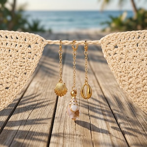 May include: Three gold-toned metal charms hanging from a beige crocheted string. The charms include a scalloped shell, a central spiral seashell with white and tan tones, and a small cowrie shell, each attached to a delicate gold-toned chain.