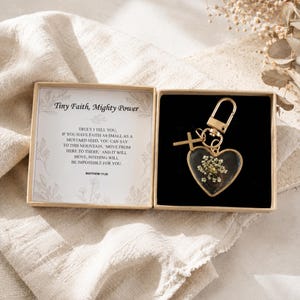 Op de afbeelding: Een goudkleurige sleutelhanger met een hartvormige hanger met een gedroogde witte bloem en een klein kruisje. De sleutelhanger zit in een open doosje met een kaartje waarop de tekst Tiny Faith, Mighty Power en Matthew 17:20 staat gedrukt.