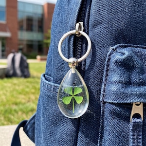 May include: A teardrop-shaped clear resin keychain containing a preserved green four-leaf clover. The keychain features a silver-toned metal split ring attached to the top, currently clipped onto the zipper pull of a dark blue denim backpack.